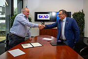 A jubilant mood fills this image — the moment after the signing of an agreement between SKAO and ESO. The two directors, smartly dressed, are standing in an office, with their pens laid down and documents on the dark brown conference table. They are leaning across and shaking hands with one another, with both smiling as they do so. On a TV monitor behind the two men, the SKAO and ESO logos are displayed. There is another screen with the blue ESO logo too, but the man on the right hand side is standing in front of it. There are also two plants on either side of the office.