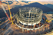 This aerial photograph shows the construction site of the Extremely Large Telescope, ELT, kissed by golden sunlight. The circular structure is made up of steel beams criss-crossing each other, all of which are sat on top of the concrete base. One gets a sense of how big the ELT structure is when comparing it to a white car, parked just left of centre at the left of the image — it is tiny compared to this colossal structure. In the bottom right small ant-like creatures are strolling about — these are the construction workers, dwarfed by the sheer scale of the ELT. Large cranes and other construction equipment are strewn around the outer edges of the structure. Off into the distance behind the construction site the brown, empty landscape with hills and smooth mountains of the Atacama desert stretches out.