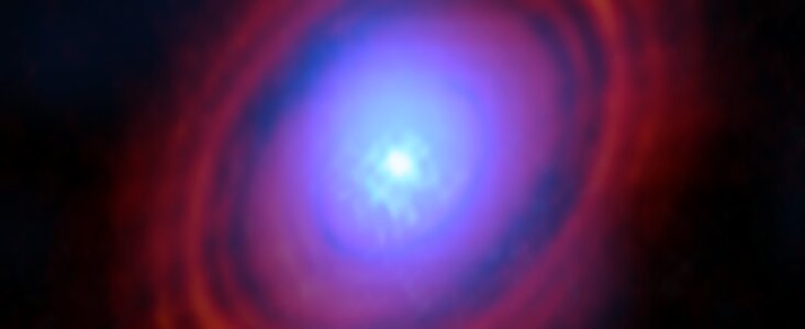 On a dark background, a bright bluish-white object at the centre of the image is surrounded by reddish rings in a disc shape. The object and rings are hazy and slightly blend.