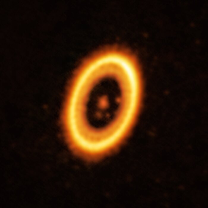 The majority of the image is black, but at its centre is the bright, glowing stellar system. A large bright orange elliptical ring, like a stretched oval donut, dominates the image. A large fuzzy orange blob is located at the centre of the ring, where the star of the PDS 70 system resides. Some smaller and fainter orange blobs, indicating planets or possible planets, orbit it.