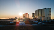 This image shows the platform on the top of Paranal mountain with the telescopes that make up ESO’s Very Large Telescope. In the background we see clouds surrounding the Paranal mountain, and the setting Sun coming down in the picture’s centre. On the right of the image, three large cylindrical structures of the Unit Telescopes can be seen. While on the left four spherical auxiliary telescopes are positioned.