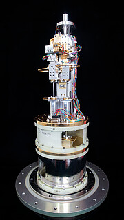 On a perfectly black background, this image shows the cold cartridge assembly of an ALMA Band 2 receiver. On top of a grey, metallic circular base, the different parts of this component are stacked on top of one another. First, a black cylinder, then white and gold ones. On top of those are many grey and gold parts, with colorful wires.