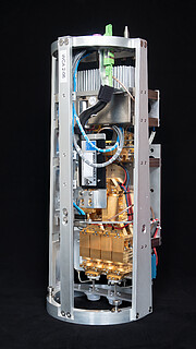 On a perfectly black background, this image shows the warm cartridge assembly for an ALMA Band 2 receiver. The cartridge is long and cylindrical in shape, with an outer structure that is predominantly grey in colour. Inside the casing, various wires and gold metallic parts are assembled.
