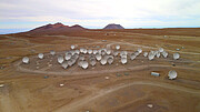 Under a cloudy and whitish sky, a light caramel plateau spans across almost all of the image. The main characters of this aerial picture are ALMA’s white antennas, closely packed together on the plateau. The photo is taken from the front of the antennas, which are pointing towards the camera, like a group of friends randomly posing. Further away, there are some dark brown mountains closely packed together on the left. Meanwhile, on the right, an isolated massive mountain stands alone.