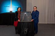 Two people stand holding a large print image of a nebula. In the background there is a large white curtain to the right and a bright blue sign to the left that says: Aniversario ESO y Chile.