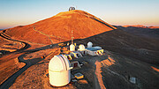 A large, rusty red, sunlit hill dominates the top half of this image. Set below it in the foreground are several white telescope domes and buildings. In the background, atop the hill, is another grey, criss-crossed telescope dome. A large shadow is cast by the hill on the right of the image, where other hills peak into the pastel horizon.