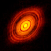 Hazy thick rings in a disc shape glow in contrast to a dark background. The rings are red near the edges and shift to shades of orange, then yellow, moving inwards.