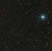 The image shows a dark area of night sky, speckled only lightly with the white and blue glow of stars. Towards the upper right region of the image there is a large bright blue star which stands out among the others.