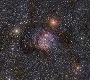 This image is dotted with countless stars that appear as shiny white dots of varying size and brightness against the black background of space. In the centre of the image, there are more stars immersed in a violet cloud. Surrounding this is the Serpens nebula, which in this image appears as a faint, dark orange glow. Several stars are still visible through the cloud though.