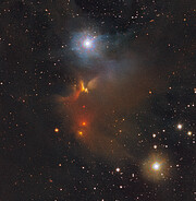 This image shows stars and clouds of gas and dust distributed over a dark background. A prominent cloud of gas and dust can be seen in the central part of the image. It features an amorphous cloud in a red and brown hue. A blue star shines brightly in the upper part, and a yellow star shines brightly in the lower right part of the image.