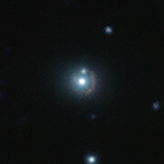 Located centrally on a dark background is a bright white circular fat round dot, which is the nearby galaxy, surrounded by a very faint reddish arc, which is the more distant 9io9 galaxy. There are a few other white blobs scattered around the image too; a couple are quite bright, while two more are fainter.