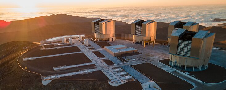 This image shows the Paranal plaftorm with the telescopes that make up ESO’s Very Large Telescope atop it. In the background we see clouds surrounding the Paranal mountain, and the setting sun on the left.