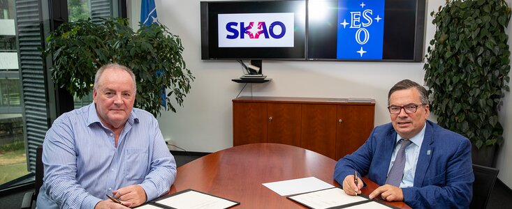 The image shows the two directors, smartly dressed, sitting down at a dark brown conference table in an office setting. Both are sitting, looking at the camera, with documents in black dossiers on the table, pens in hand, ready to sign the cooperation agreement. On two TV monitors behind the gentlemen, the logos of the respective organisations are displayed — SKAO on the left and ESO on the right. There are also two plants on either side of the office.