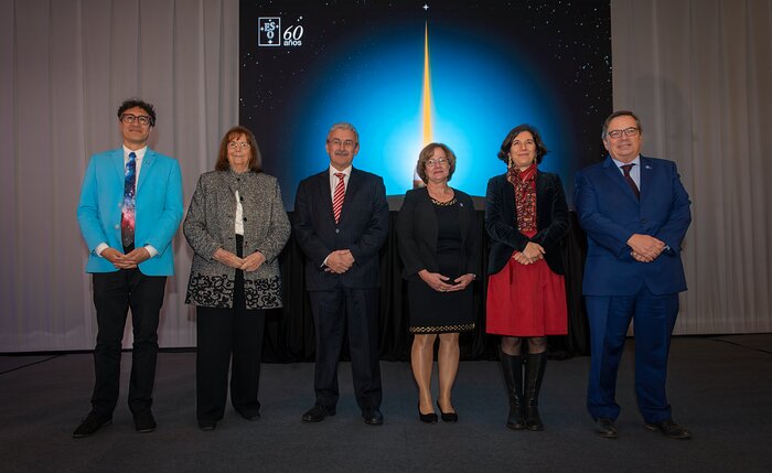 Sechs elegant gekleidete Personen stehen in einer Reihe vor einem großen Bild des Himmels mit dem ESO-Logo und den Worten 60 anos