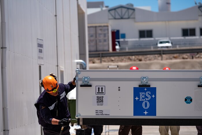Outside a white building to the left of the photo, a person in construction clothing and a protective helmet is working on a large white box with the ESO logo — four stars around the letters E, S, O on a blue background — on it. In the blurry background is another white building.
