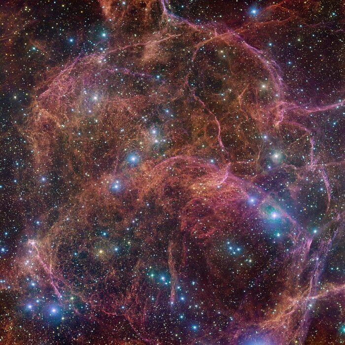 The image shows stringy clouds of hot pink and golden orange, nested together all over the frame, almost like the messy fragments of a spider’s web. Close to these colourful clouds, we find bright blue massive stars. In between the clouds there are gaps, revealing stars in blue and yellow, almost like sparklers.