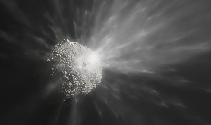 This greyscale image shows an asteroid with a rocky texture over a black background. In the centre of the asteroid there is an impact crater. Streaks of ejected debris emanate from the crater, forming a filamentary halo of debris around the asteroid.