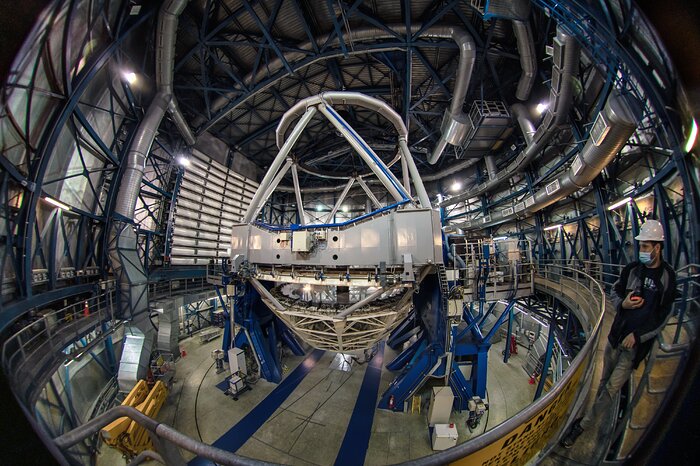 The blue and metallic structure of the third Very Large Telescope is placed in the middle of this image. The telescope is pointing upwards, enclosed in its dome of similar blue and metallic steel construction. The image is taking with a fisheye lens, in order to capture the size of the large telescope. To the right, a person stands on a platform that goes around the whole interior of the dome, looking towards the telescope.