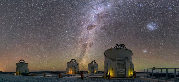 A landscape picture of the VLT's four auxiliary telescopes against the vast background of the night sky. The Milky Way, in the centre of this image, is pointing upwards and is surrounded by countless stars. The Large Magellanic Clouds are also visible. Towards the horizon in the sky, a glow of orange and green appears.