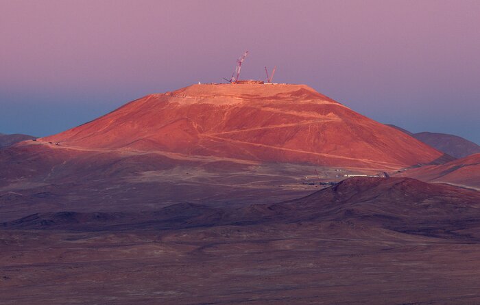 A landscape image of the desolate Atacama desert with the mountain, Cerro Armazones, in the centre at quite some distance from the photographer. The red desert is mostly in shadow, except for the top of Armazones, which is still illuminated by the setting sun. The service road and cranes are clearly visible and the only markers of human life. The sky is a beautiful pink that fades into dark blue towards the horizon.