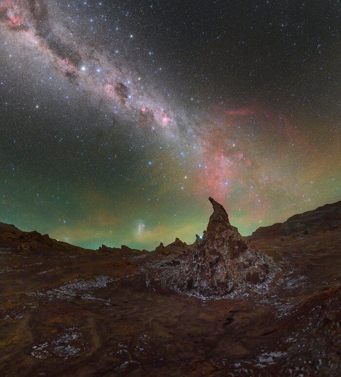 The whole image feels alien to look at. The upper two thirds constitute the night sky, a dark canvas painted with thousands of stars, the hazy white band of the Milky Way stretching off to the upper left, and wispy green and red features clinging near the horizon. The bottom third of the image curves like a bowl as the desert floor stretches up in front of the lens. Pointed features poke out of the dusty red ground, which is speckled with white flecks of salt.